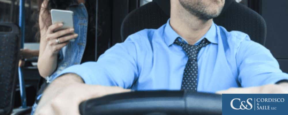 A lady standing behind a bus driver with her smart phone